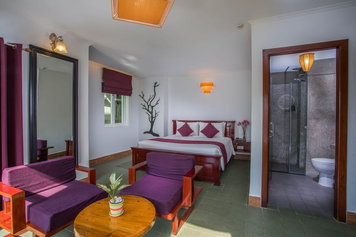A hotel room with a bed, a sofa, a chair, a small table with a plant, a mirror, a wall-mounted light, and a shower area.