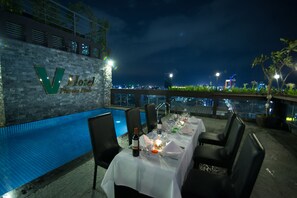A rooftop dining area with a pool, a table set for two, and a view of the city skyline at night.