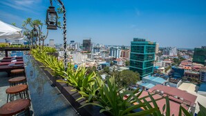 A rooftop bar with a city view, featuring a long counter with stools, potted plants, and hanging lanterns. - V Hotel Phnom Penh (Phnom Penh)