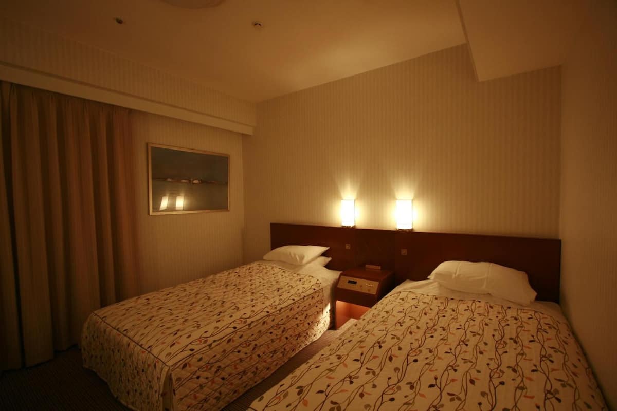 Habitación de hotel con dos camas, una mesita de noche, una lámpara y una ventana con cortinas.