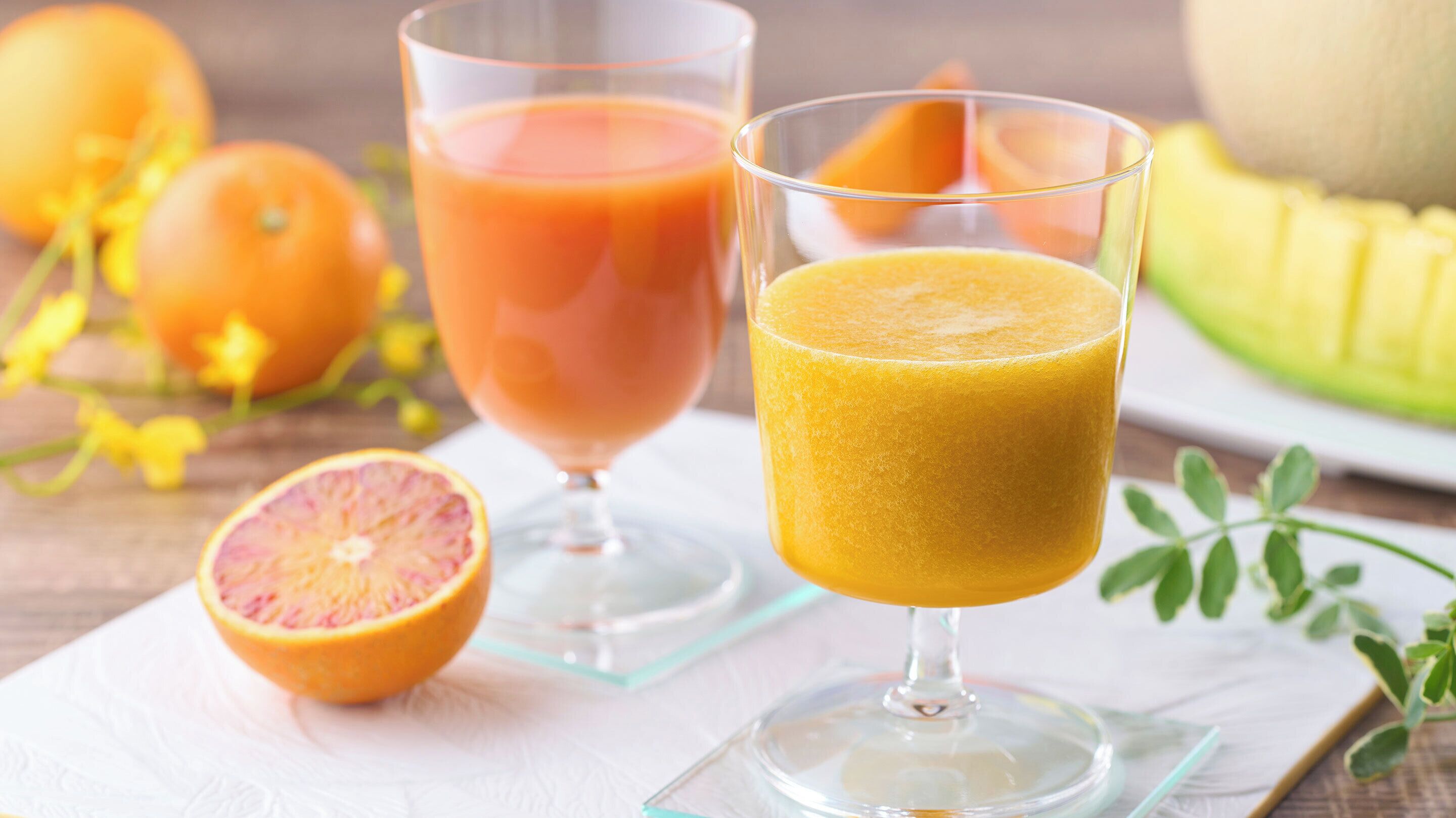 Deux verres de jus, l’un contenant un liquide orangé rosé et l’autre un liquide orangé jaunâtre, posés sur une serviette blanche, avec un fruit coupé en deux et des fruits entiers en arrière-plan.