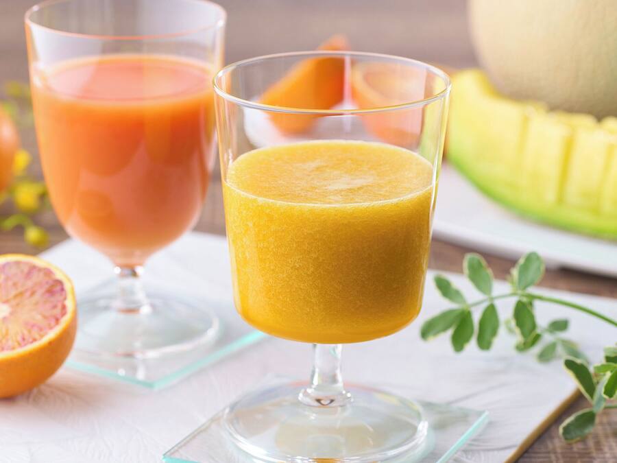 Dos vasos de jugo, uno con líquido rosado-anaranjado y otro con líquido amarillo-anaranjado, sobre una servilleta blanca con una fruta cortada por la mitad y frutas enteras al fondo.