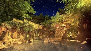 A stone-tiled hot spring area surrounded by rocks and trees, with a bamboo fence and traditional Japanese architecture in the background. - Ooedo Onsen Monogatari Miyoshiya (Shinonsen)