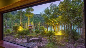 A view through a window of a garden with a stone lantern, bamboo fence, and a body of water. - Ooedo Onsen Monogatari Miyoshiya (Shinonsen)