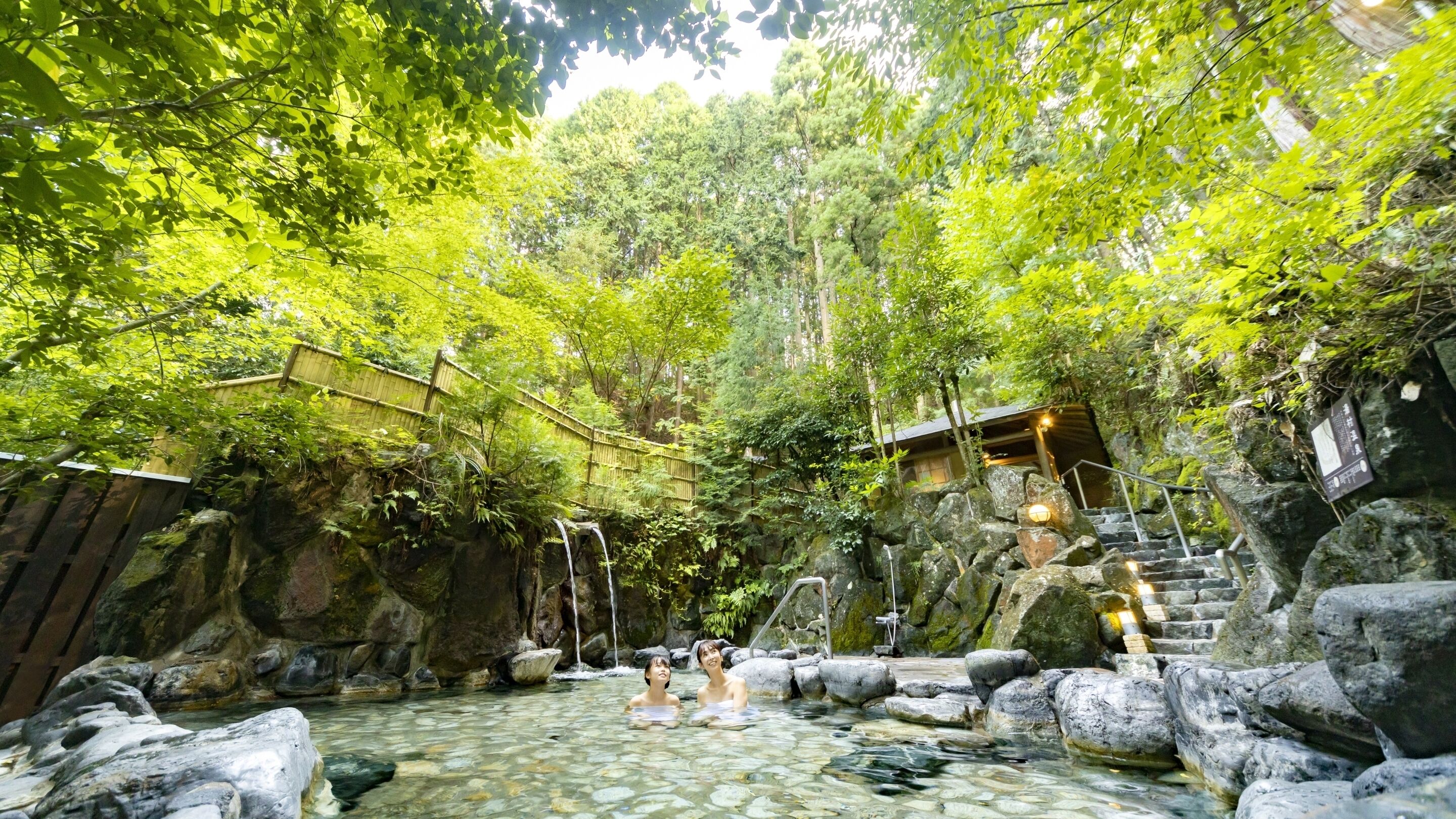 A natural hot spring surrounded by rocks and trees, with a wooden deck and stairs leading up to a building.