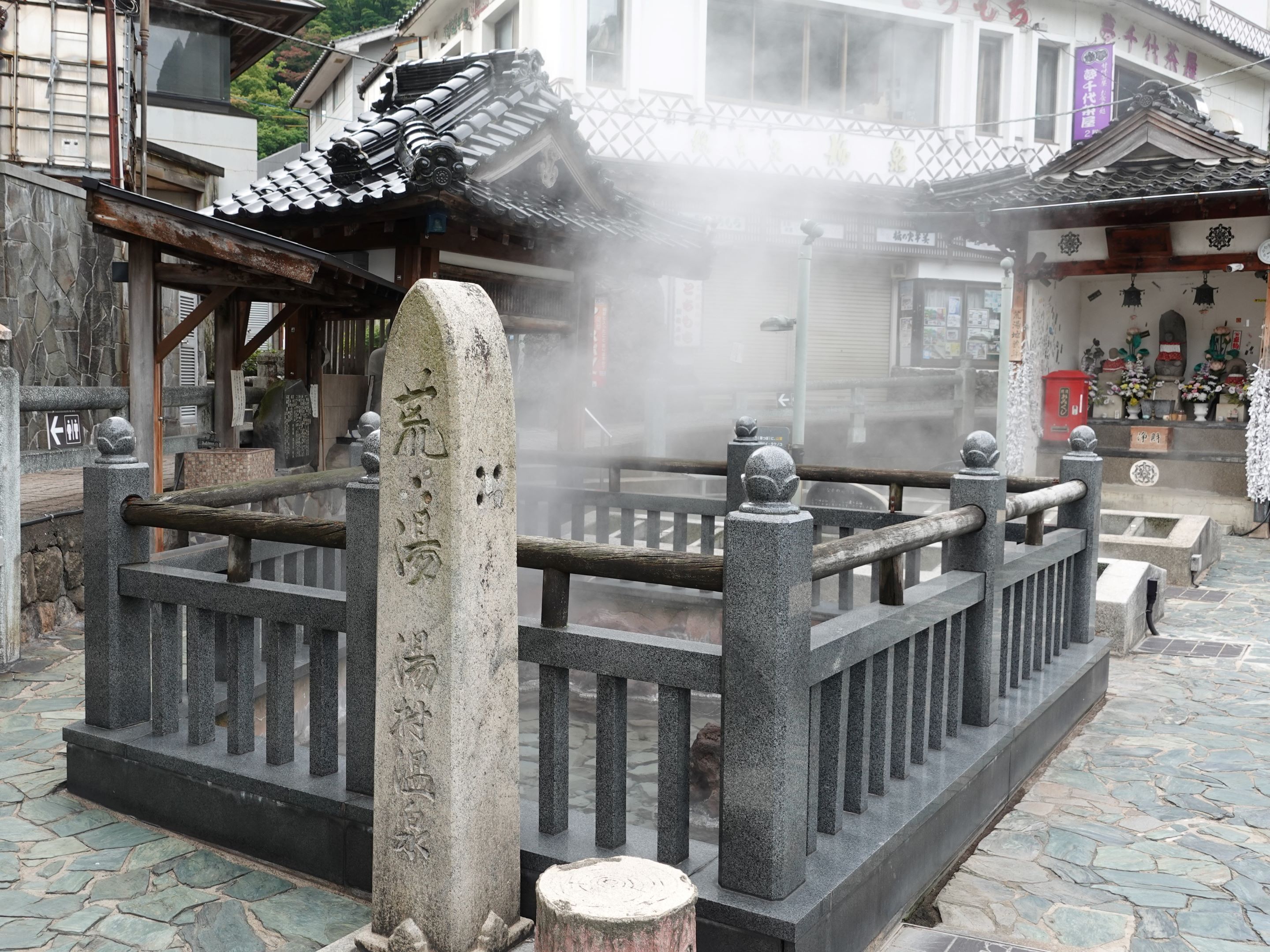 Un portón japonés tradicional con pilares de piedra y techo de tejas, que da a un patio con suelo de piedra.