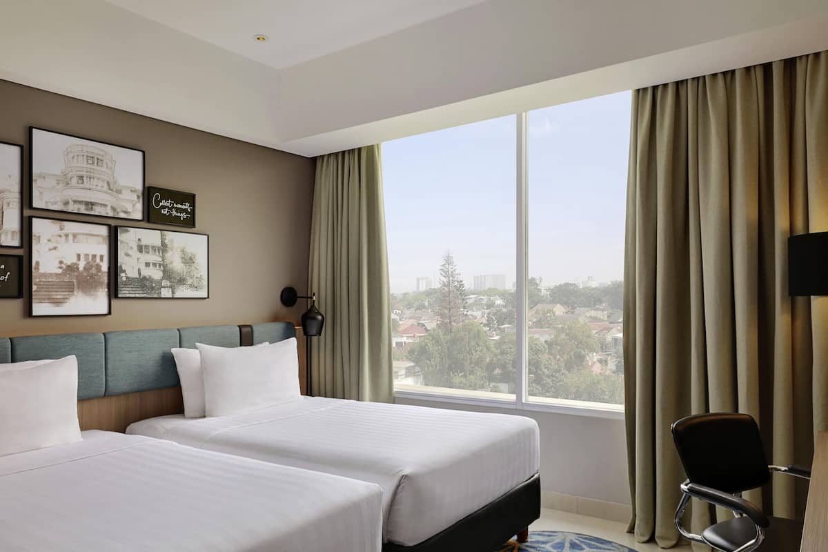 Ein Hotelzimmer mit zwei Betten, einem Schreibtisch, einem Sessel und einem großen Fenster mit Vorhängen.