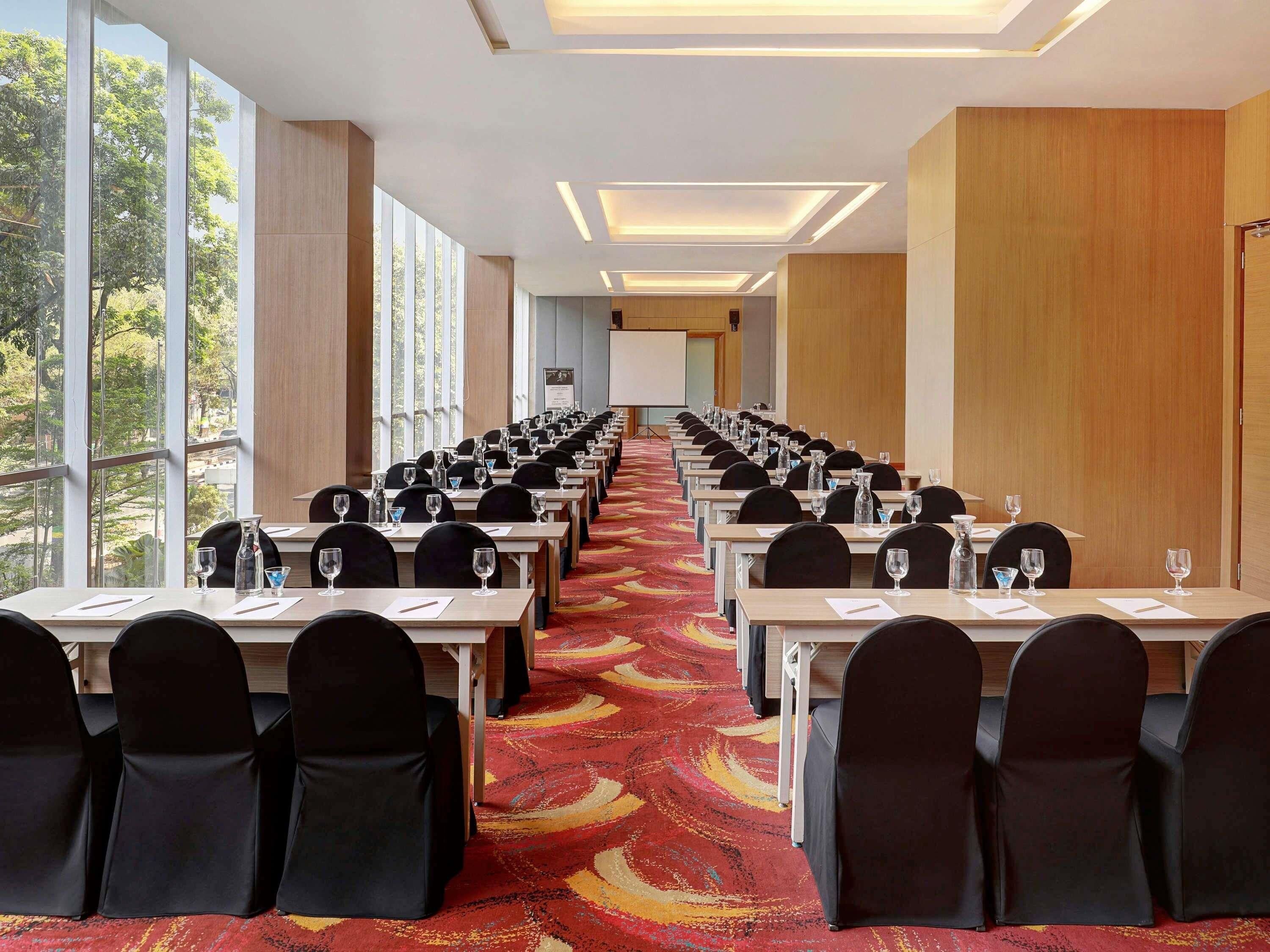 Une salle de conférence avec des rangées de tables et de chaises, de grandes fenêtres et un sol recouvert de moquette.