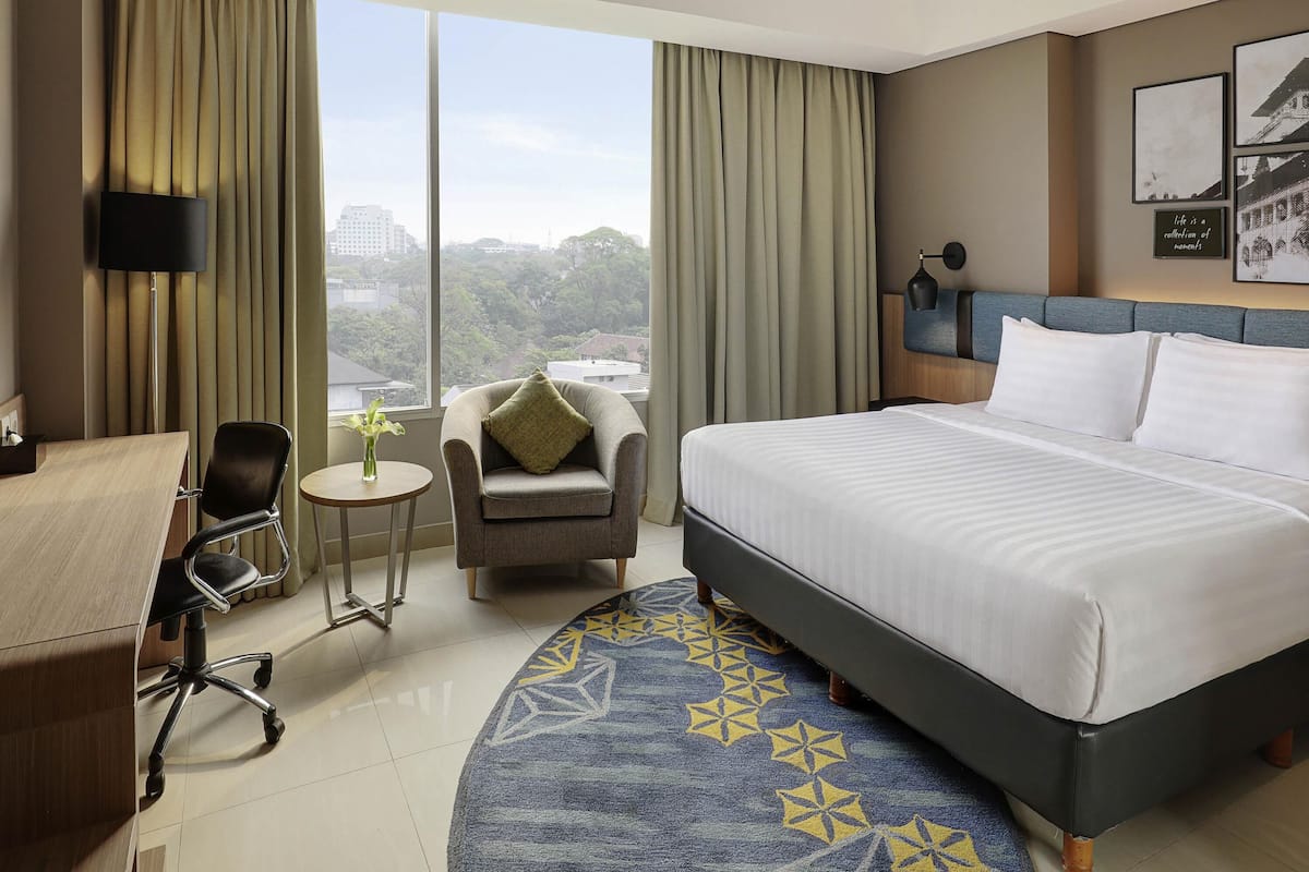 A hotel room with a bed, a desk, a chair, a small table with a vase, and a view of the city.