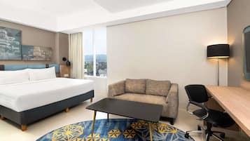 Habitación de hotel con cama, sofá, escritorio, silla, lámpara y vistas de la ciudad a través del ventanal.