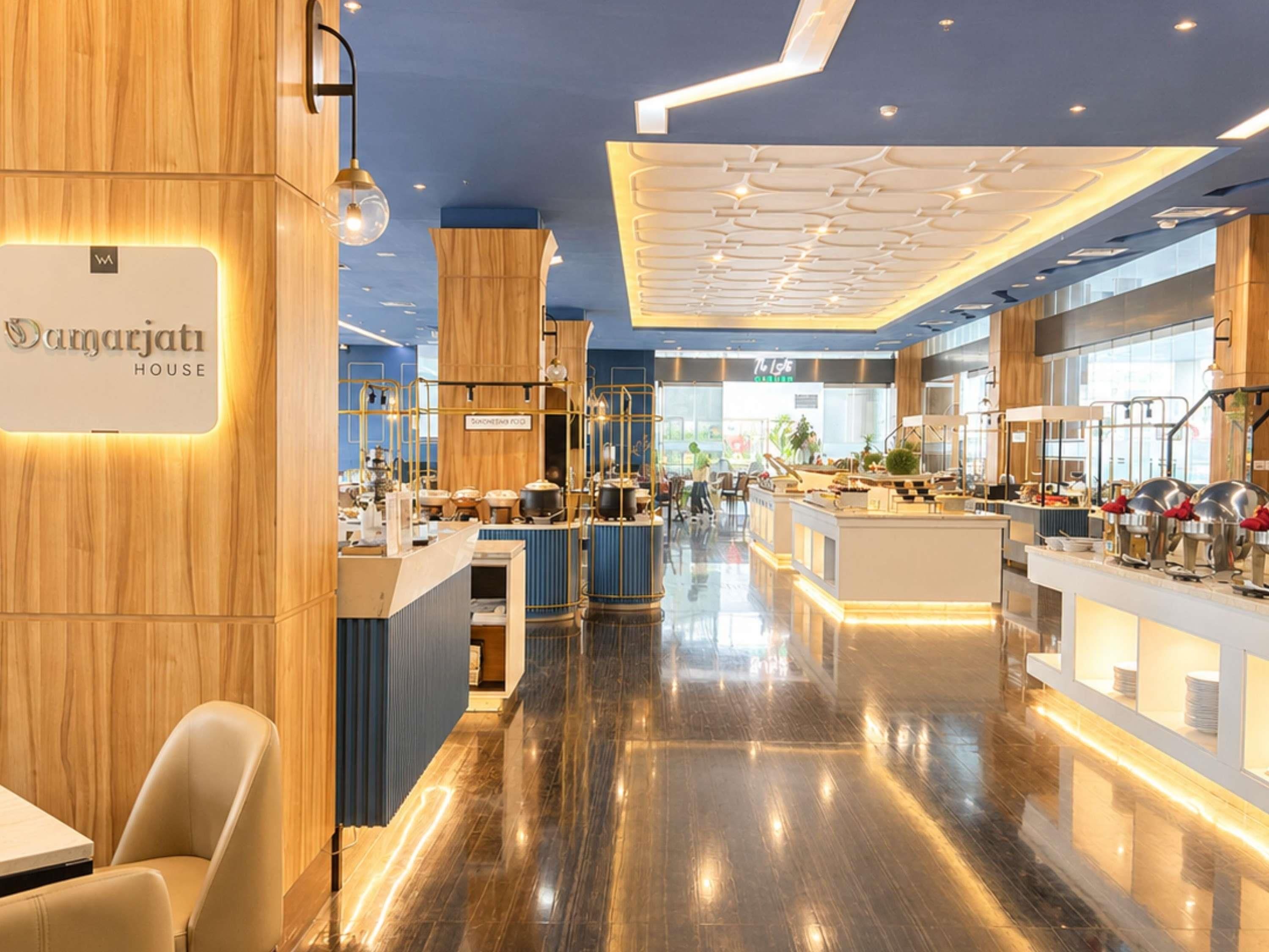 A modern hotel interior with a central reception desk, a dining area, and a kitchen visible in the background.