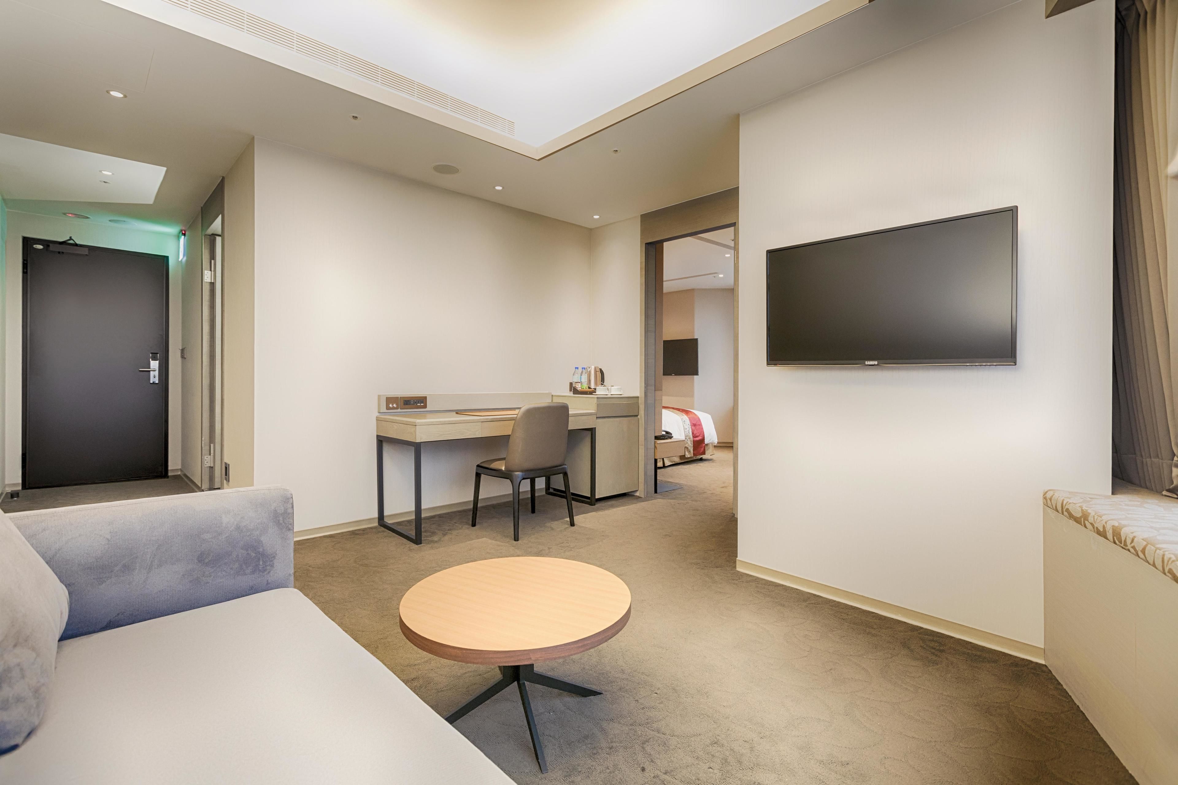 Une chambre d’hôtel comprenant un canapé, une table ronde, un bureau avec une chaise, un téléviseur fixé au mur et une porte donnant sur une autre pièce.