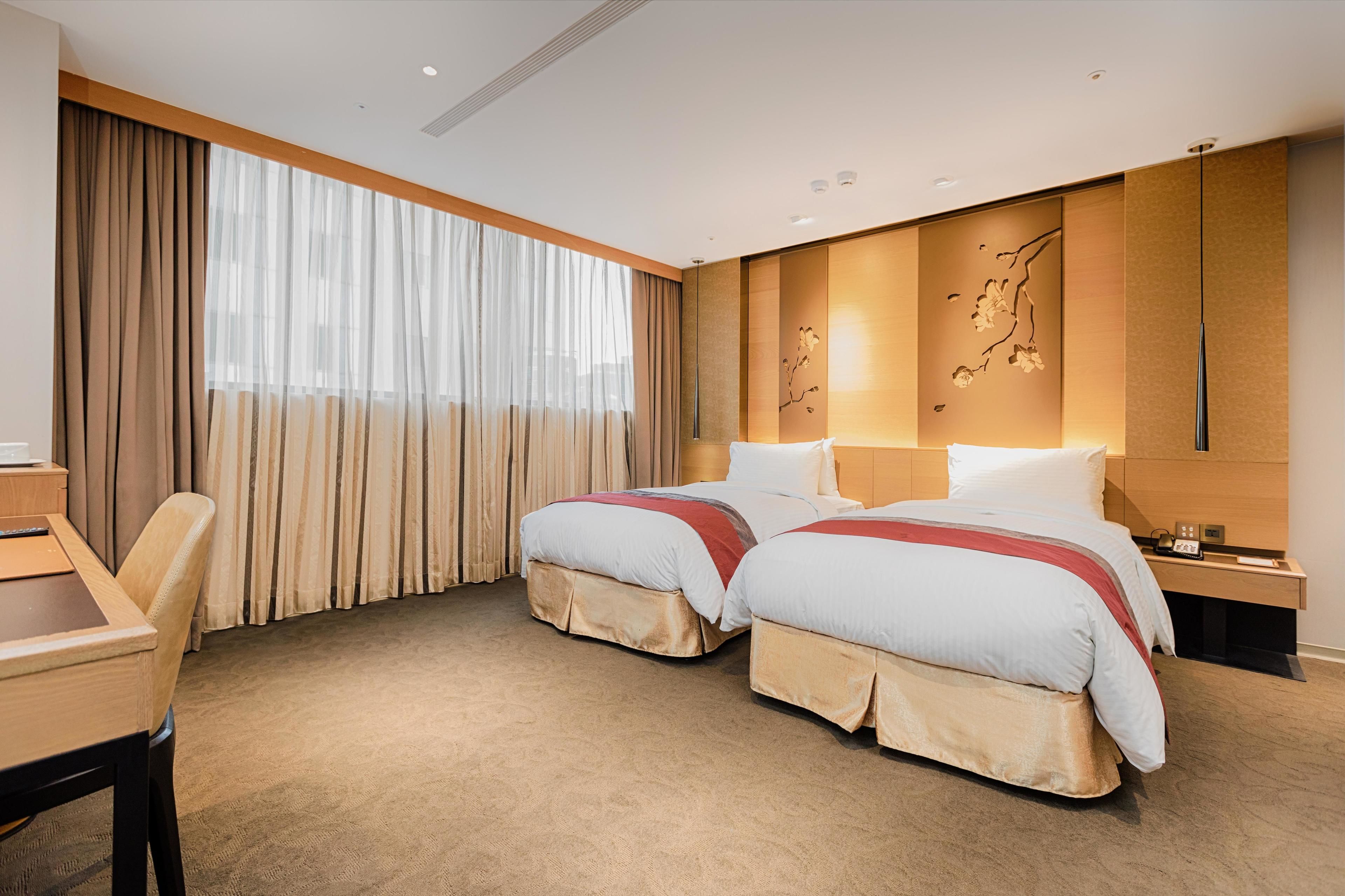 A hotel room with two beds, a desk, and a large window with curtains.