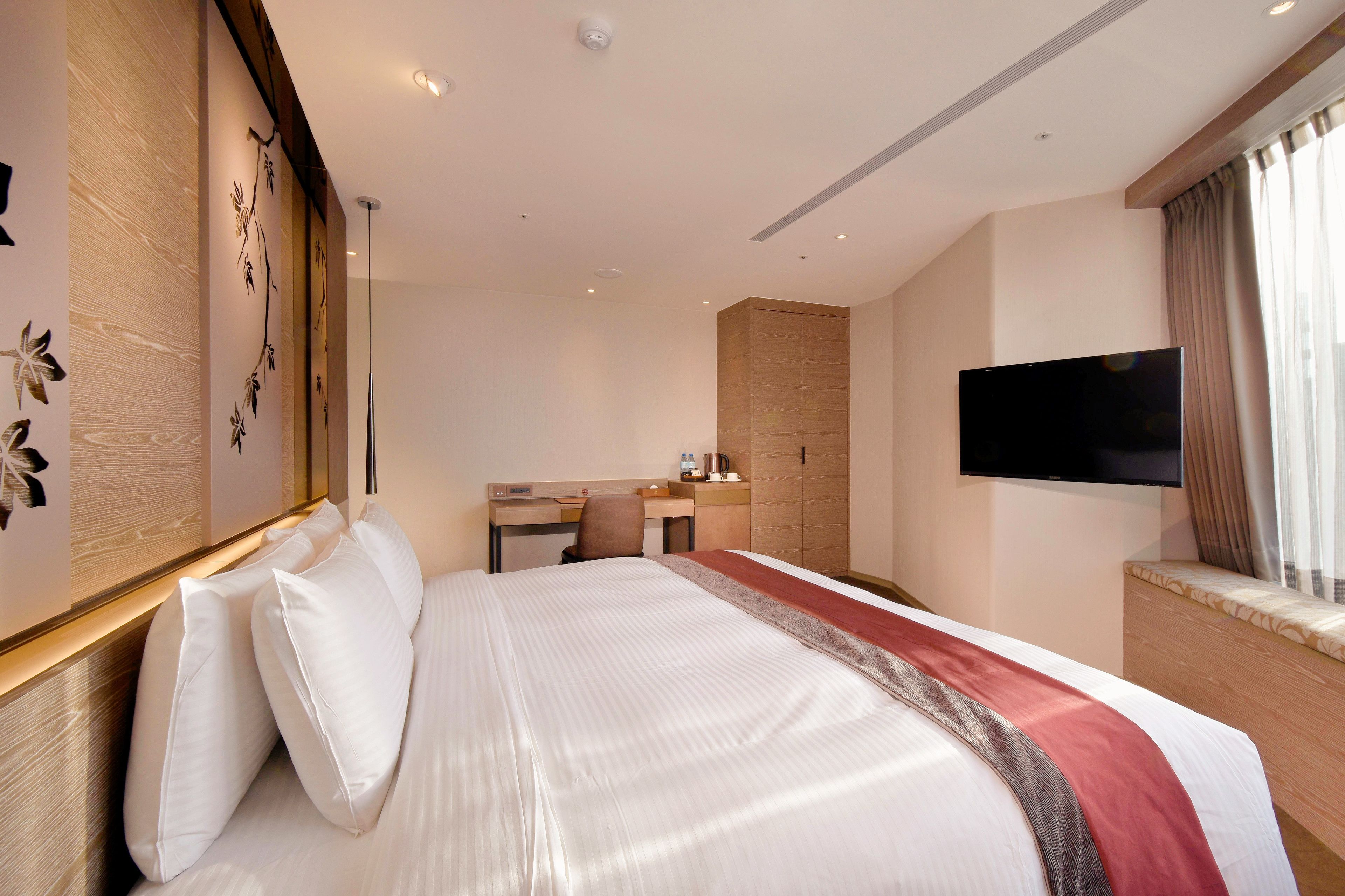 A hotel room with a large bed, a desk with a chair, a flat-screen TV mounted on the wall, and a window with curtains.