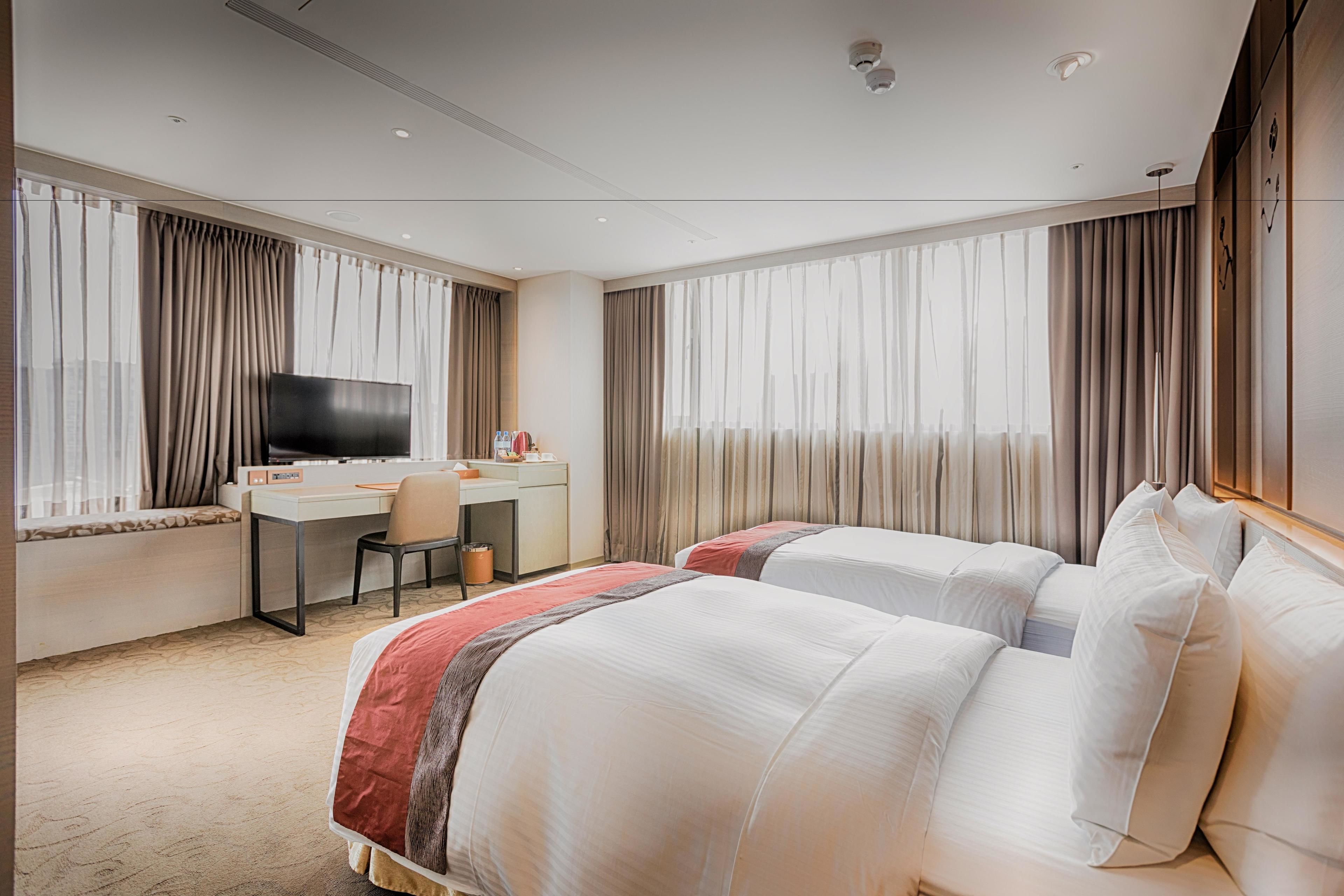 A hotel room with two beds, a desk, a chair, a television, and large windows with curtains.