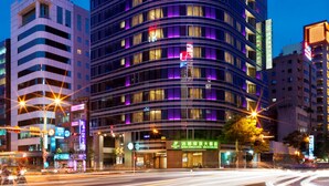 Un hotel a più piani con facciata curva, illuminato da luci viola, situato all'incrocio di una strada trafficata.