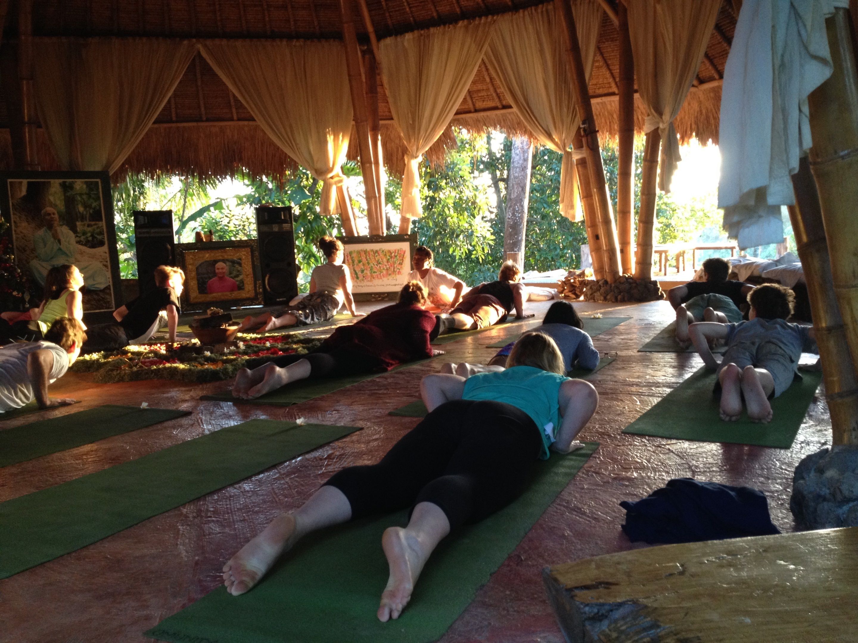 Un groupe de personnes pratiquant le yoga dans un espace extérieur abrité, doté d’un toit de chaume et de piliers en bambou.
