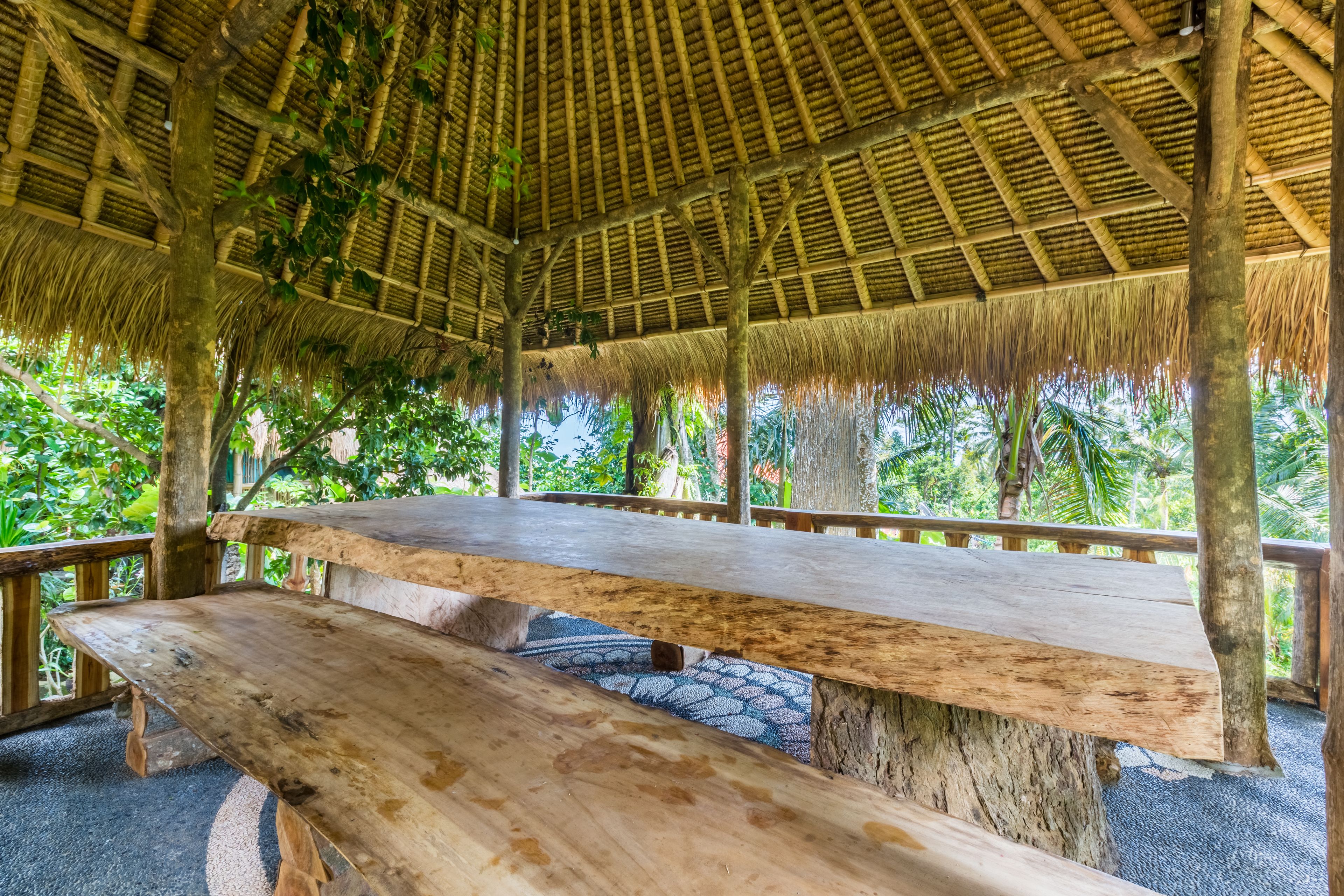 Un banc en bois, placé sous une structure à toit de chaume, entouré d’une végétation luxuriante.