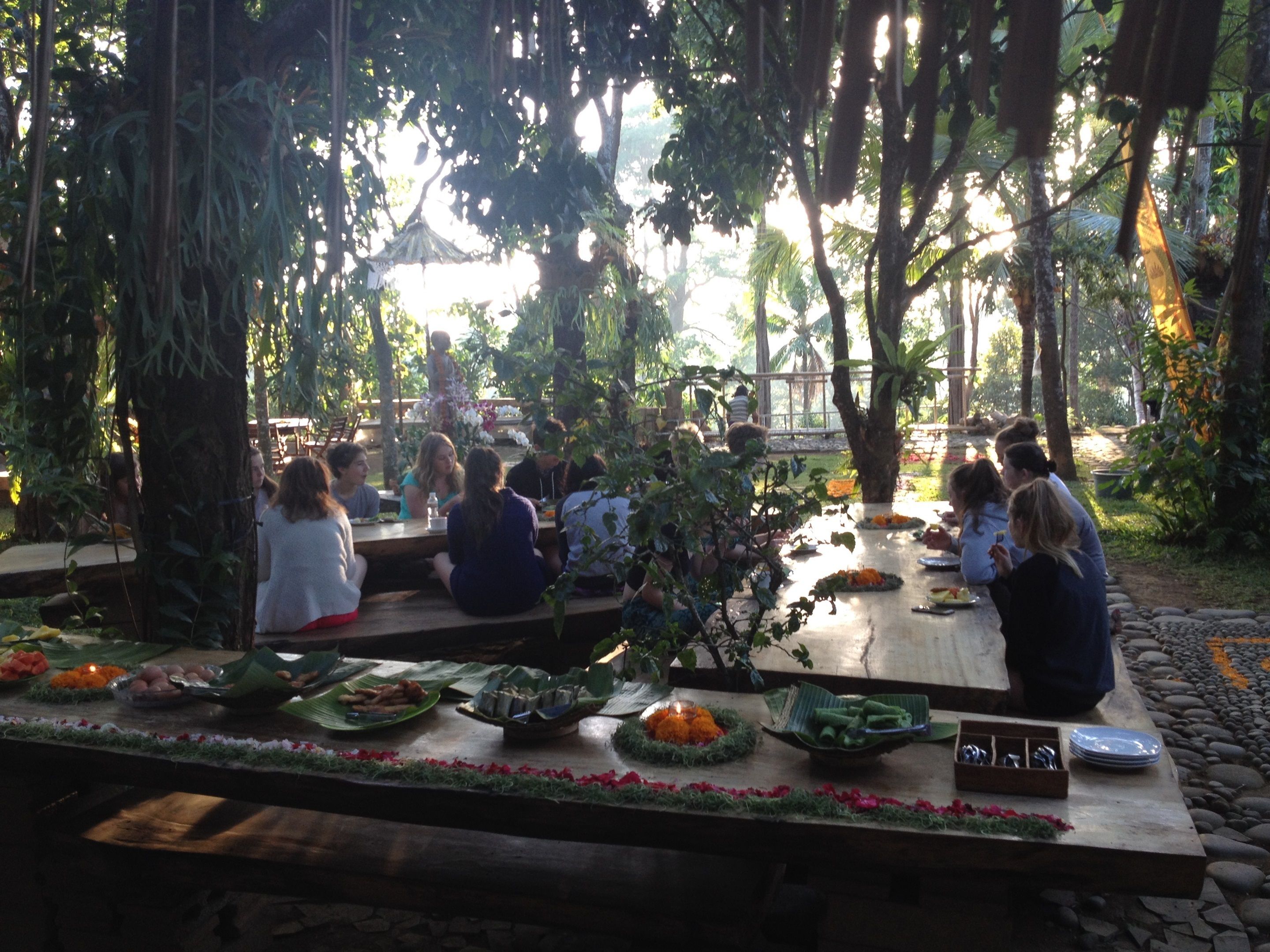 A group of people dining outdoors at a wooden table with various dishes and utensils.