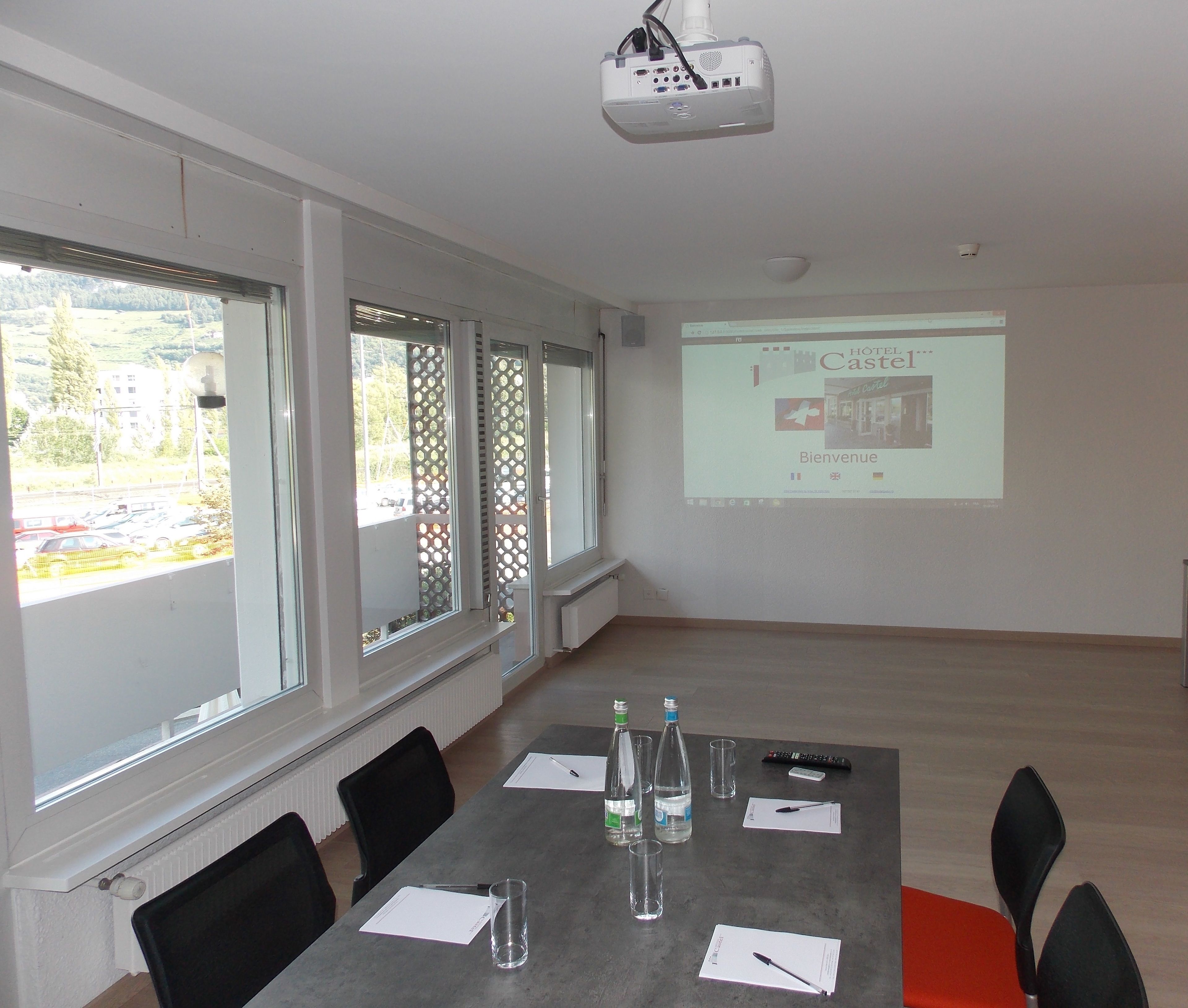 Una sala de conferencias con una pantalla de proyector mostrando una presentación de bienvenida del hotel, una mesa con documentos y vistas a un estacionamiento a través de amplios ventanales.