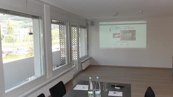 Ein Konferenzraum mit Projektionsfläche, die eine Willkommensanzeige des Hotels zeigt, einem Tisch mit Unterlagen und Blick auf einen Parkplatz durch große Fenster.