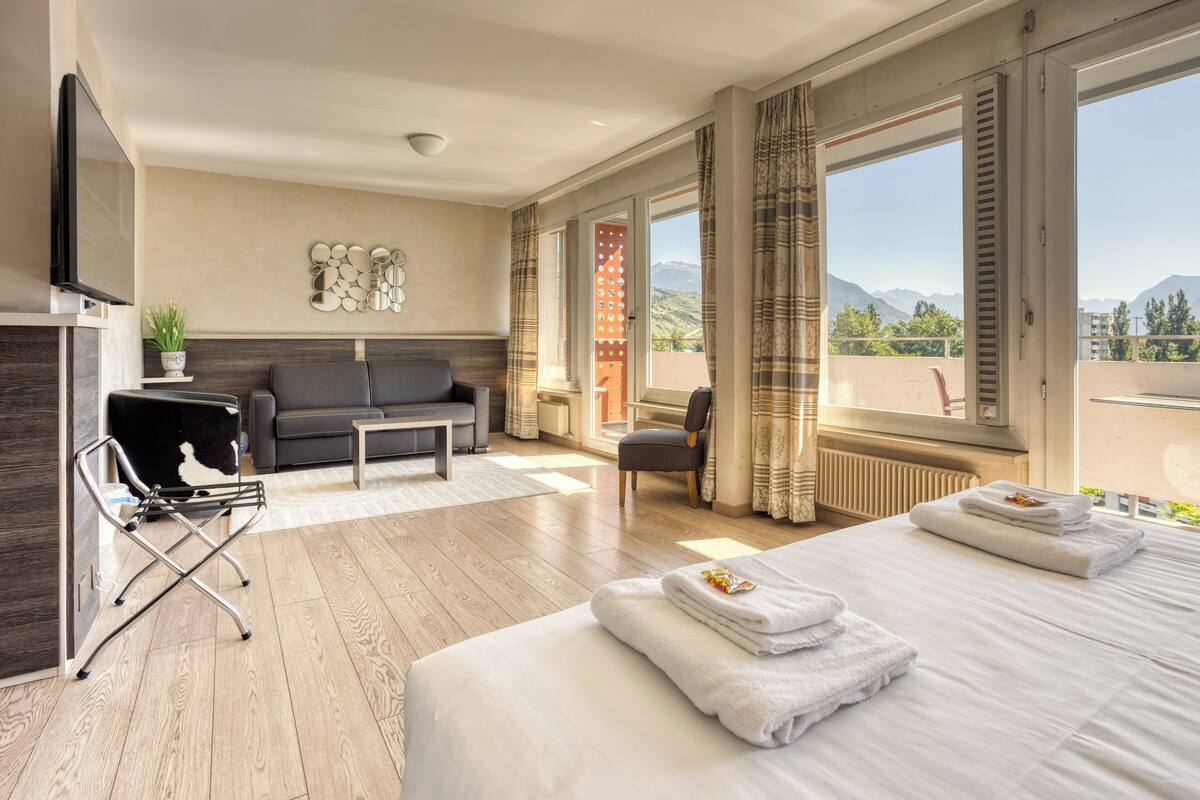 Habitación de hotel con cama, sofá, televisión y vistas a montañas.