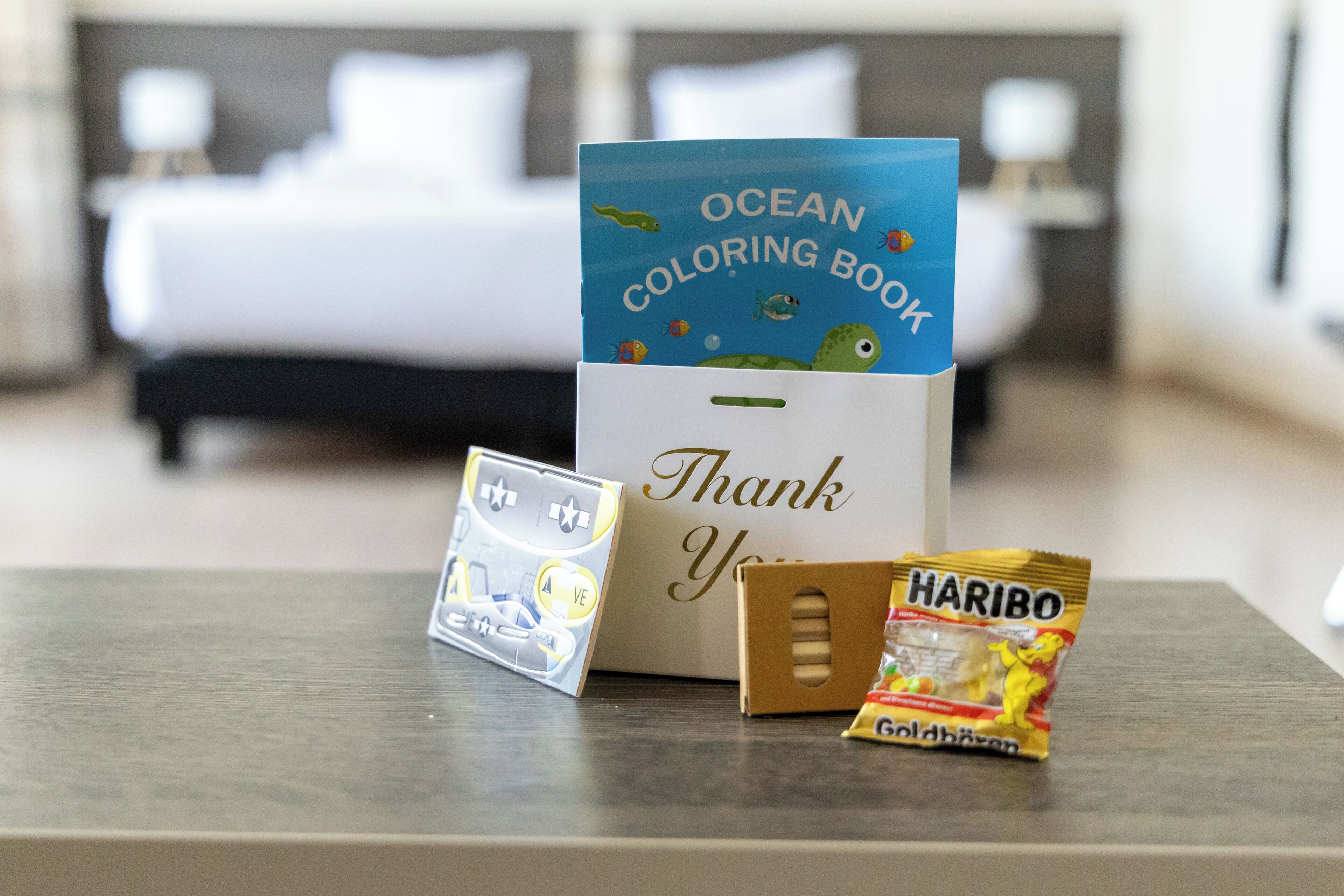A hotel room with a bed, a thank you card, a coloring book, and a snack package.