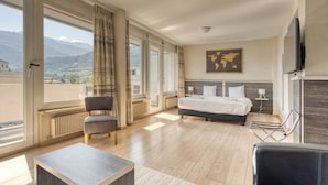 Una habitación de hotel moderna con una cama grande, una zona de estar con una silla, una mesita y vistas a las montañas a través de las ventanas.