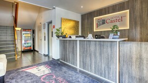 Zona de recepción de hotel con diseño moderno, que incluye un mostrador de recepción, un refrigerador de Coca-Cola y un mostrador con una planta y un letrero que dice "HOTEL CASTEL".