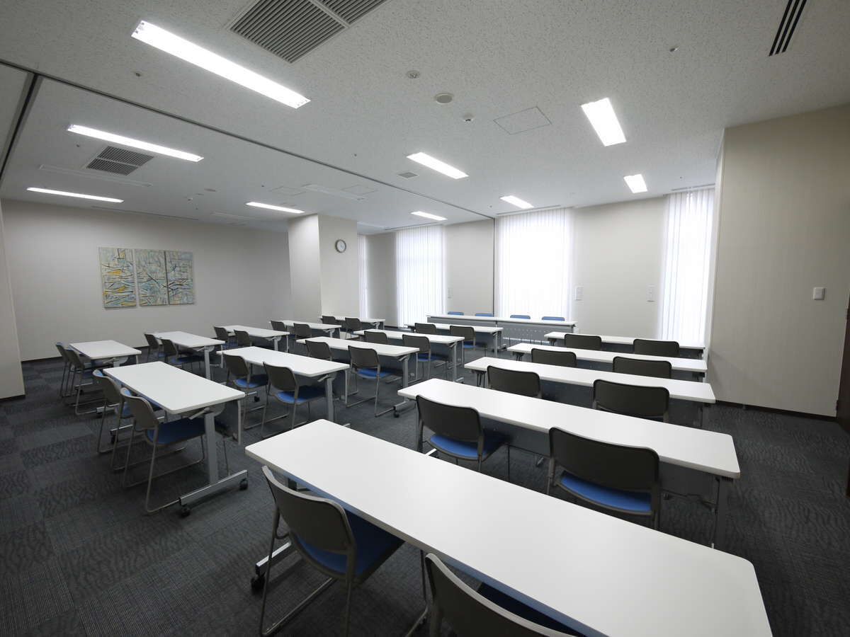 Un aula con filas de escritorios y sillas, paredes blancas y ventanas amplias.