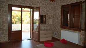 A bedroom with stone walls, wooden flooring, a bed with red towels, a dresser, and a window with a view of greenery. - Pyrgos Luxury Country Home (Rhodes)