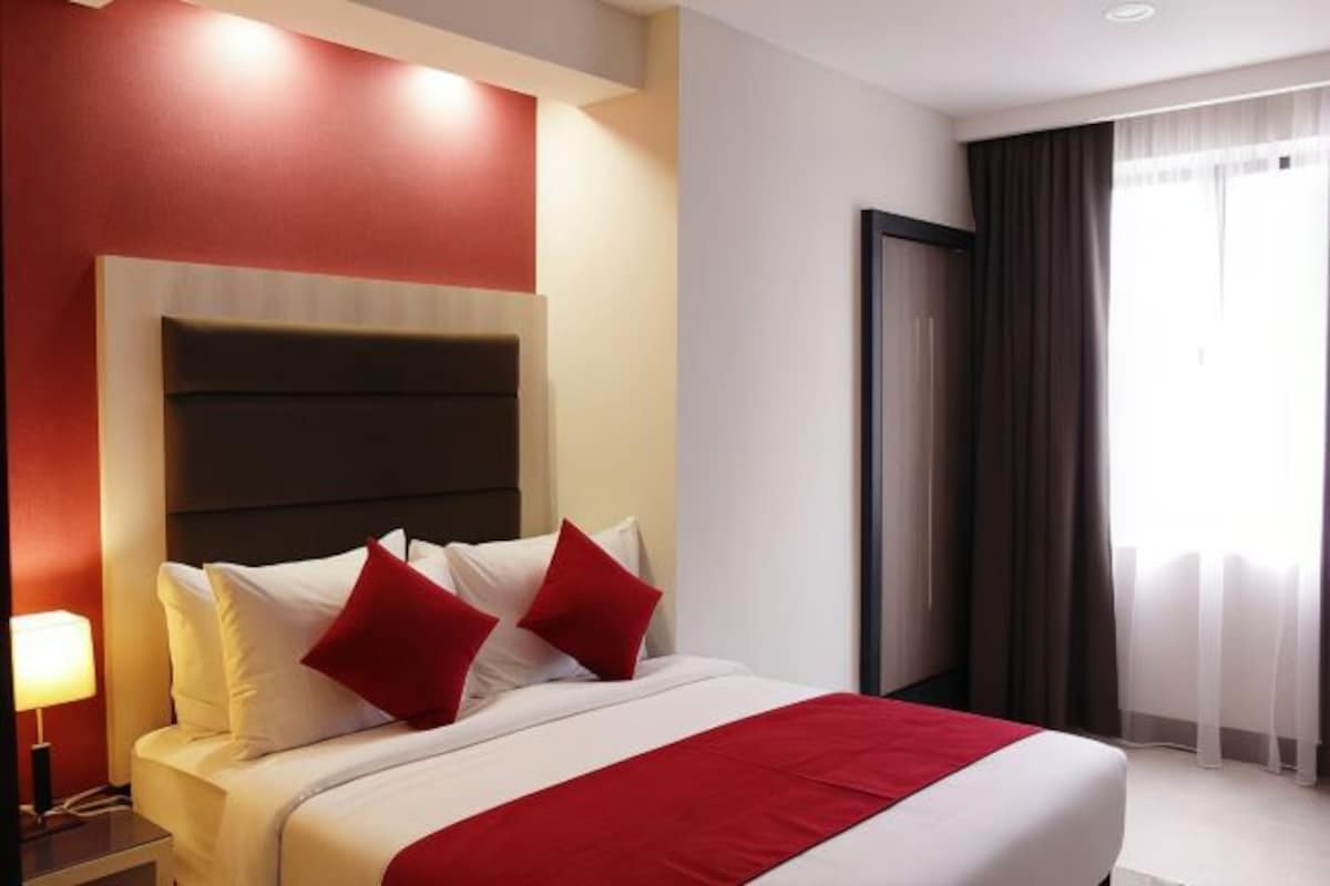 A hotel room with a large bed, red and white bedding, a nightstand with a lamp, and a view of the outdoors through a window with curtains.