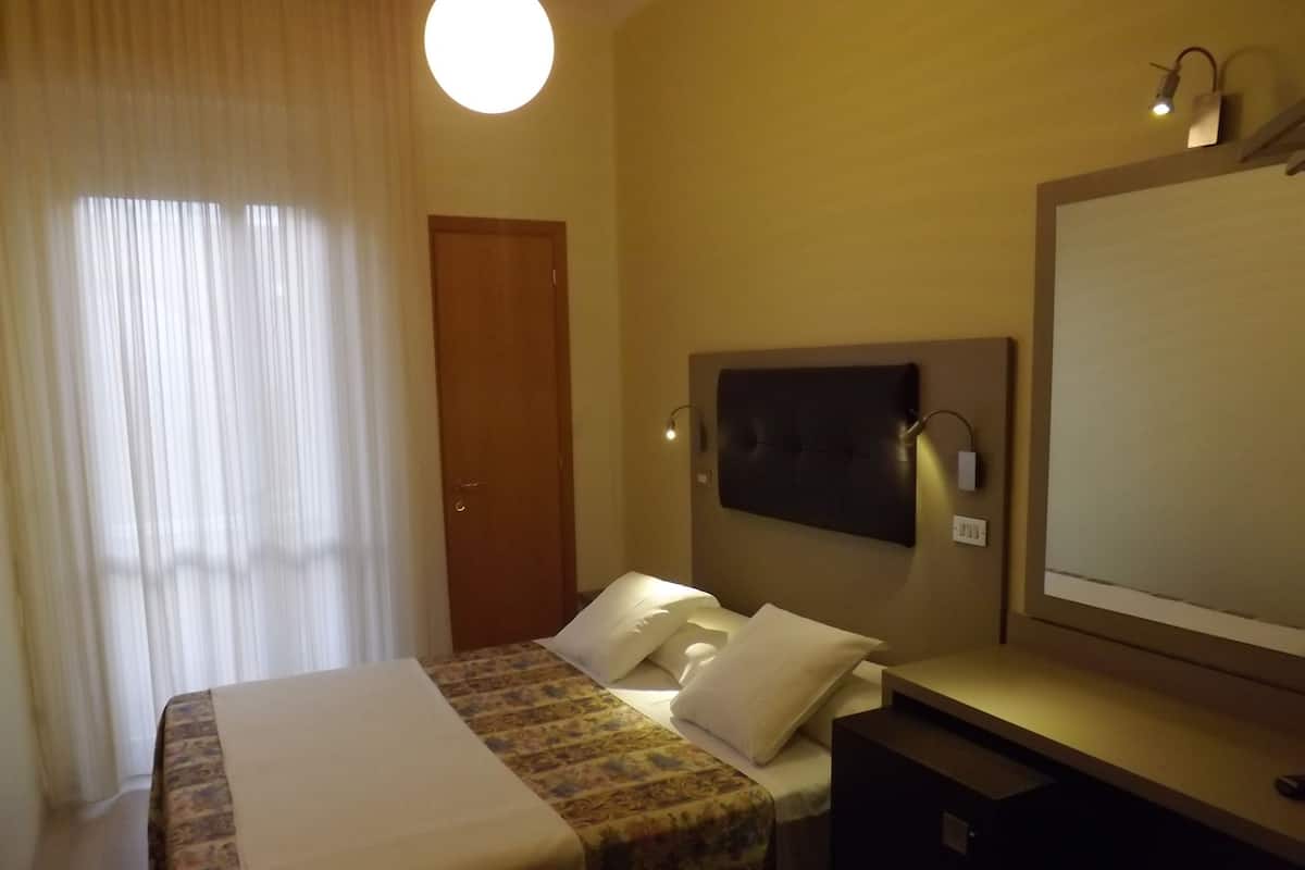A hotel room with a bed, a bedside table, a mirror, and a window with curtains.