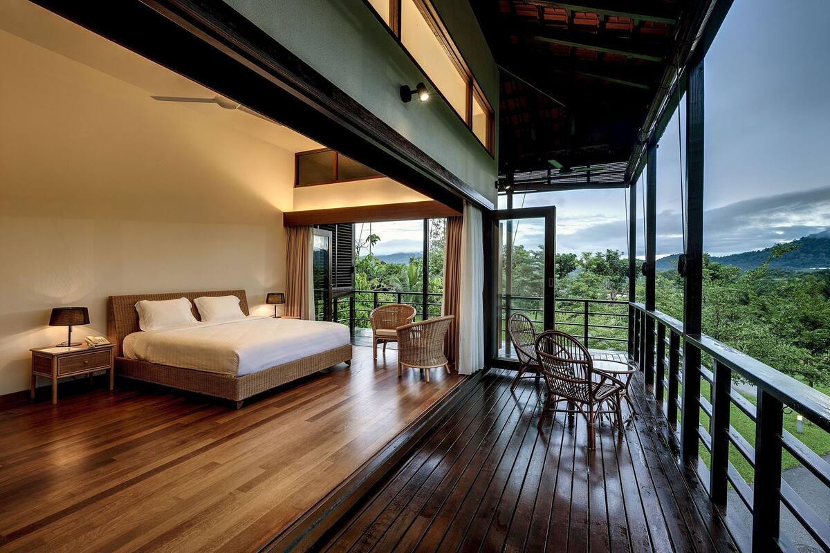 A bedroom with a bed, nightstand, lamp, and wicker chairs.