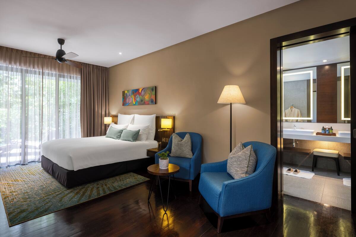 Una habitación de hotel con una cama, dos sillones, una lámpara de pie, un cuadro y un baño visible a través de una puerta de vidrio.