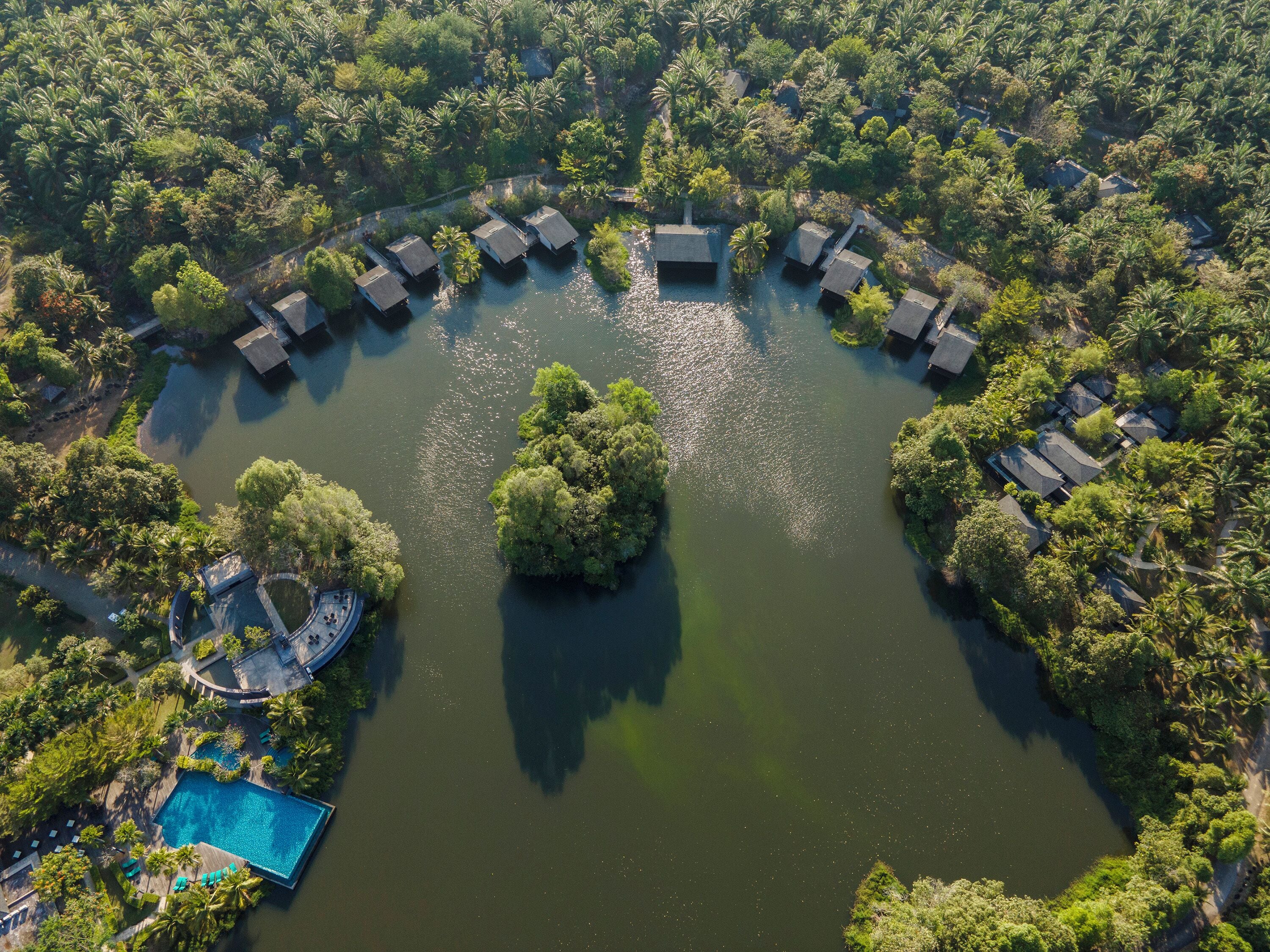 An aerial view of a resort with overwater bungalows arranged in a semi-circle around a central water body, surrounded by dense greenery.