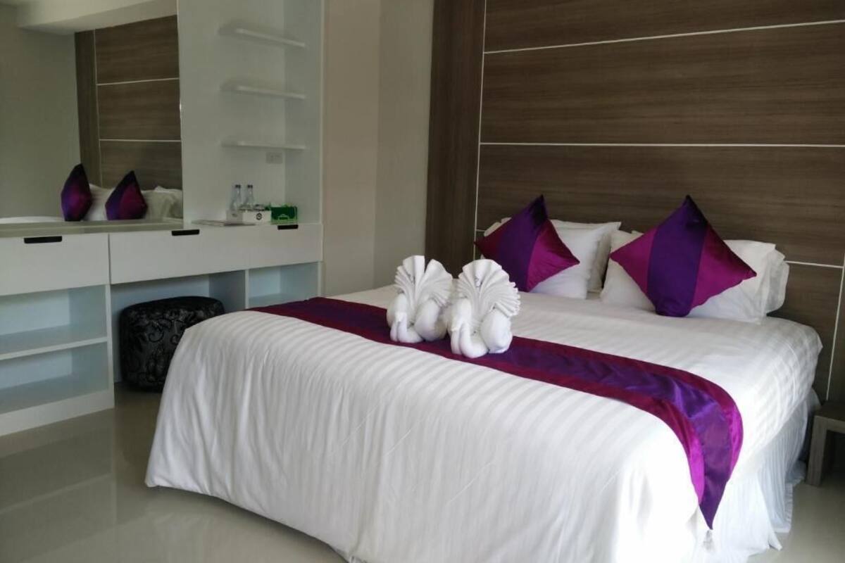 A modern hotel room with a large bed, white bedding, purple pillows, and a wooden headboard.