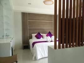 Una habitación de hotel moderna con una cama grande, ropa de cama blanca y almohadas moradas. Se observa un cabecero de madera y una pequeña mesa con una silla.