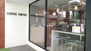 Una cafetería con fachada de vidrio, un cartel de menú visible y un espacio con taburetes.
