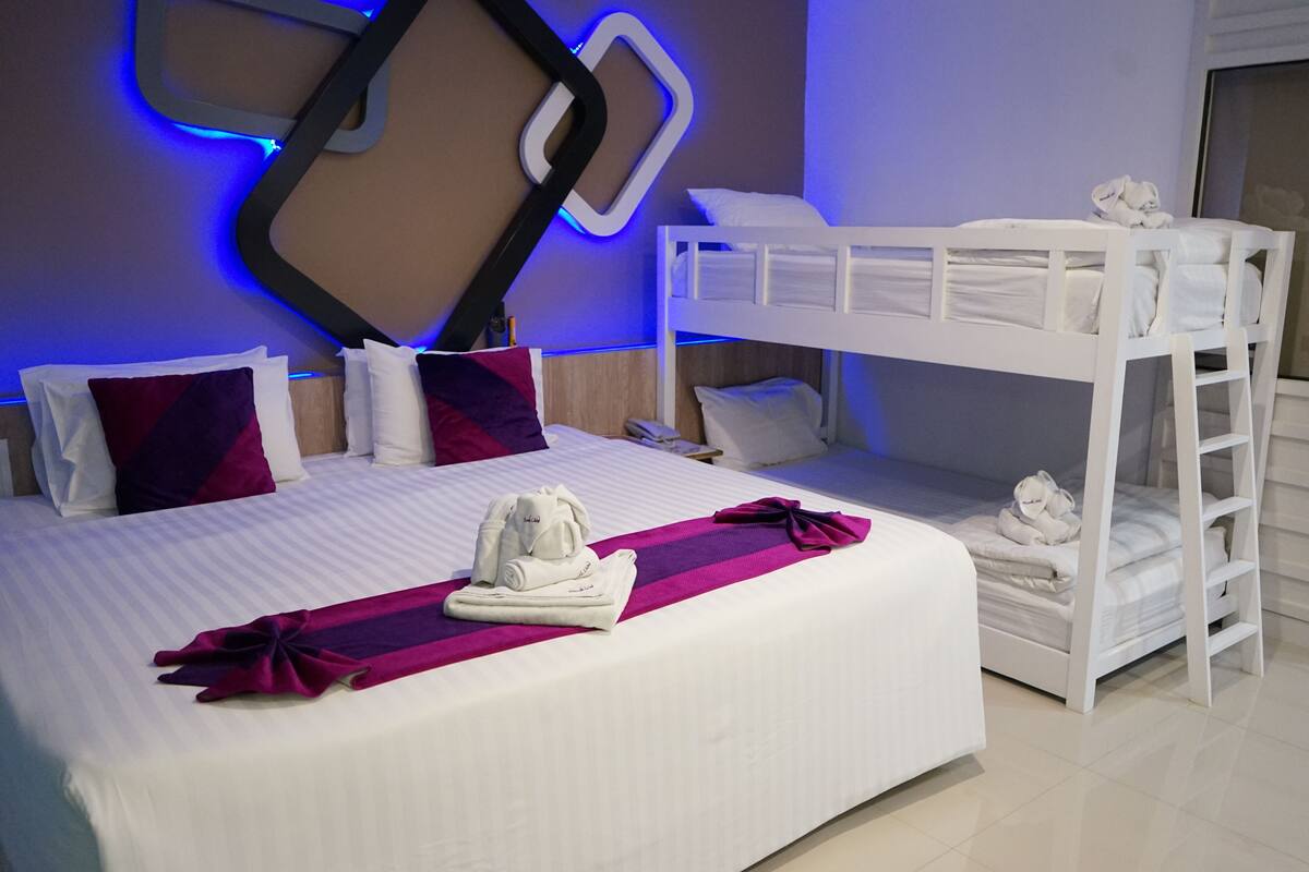 A hotel room with a bunk bed, a neatly made bed with a purple towel, and a wall-mounted light fixture.