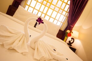 A hotel room with a bed, a window with a yellow grid pattern, and a nightstand with a lamp and flowers.