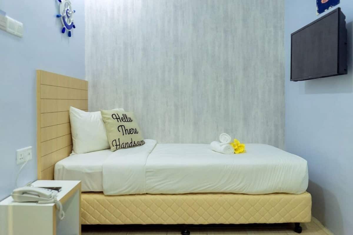 Una cama individual con colchón acolchado, una mesita de noche con un teléfono, un reloj de pared y un televisor de pantalla plana montado en la pared.