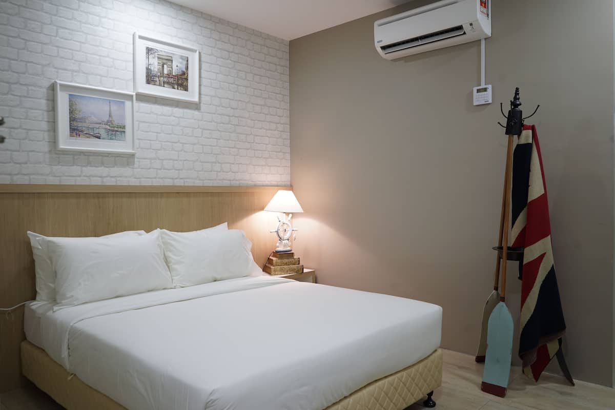 Una cama bien hecha con sábanas blancas, un cabecero de madera y un aire acondicionado instalado en la pared. Hay dos cuadros enmarcados en la pared y una paleta de madera apoyada contra ella.