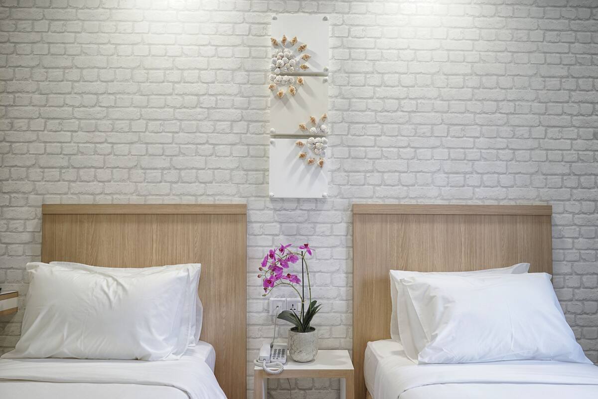 Habitación de hotel con dos camas, ropa de cama blanca, una mesita de noche pequeña con un teléfono y una orquídea en maceta, y una pared con diseño de ladrillo.