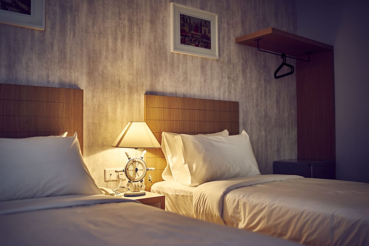 Habitación de hotel con dos camas, una mesita de noche con lámpara, un reloj y un cuadro en la pared.