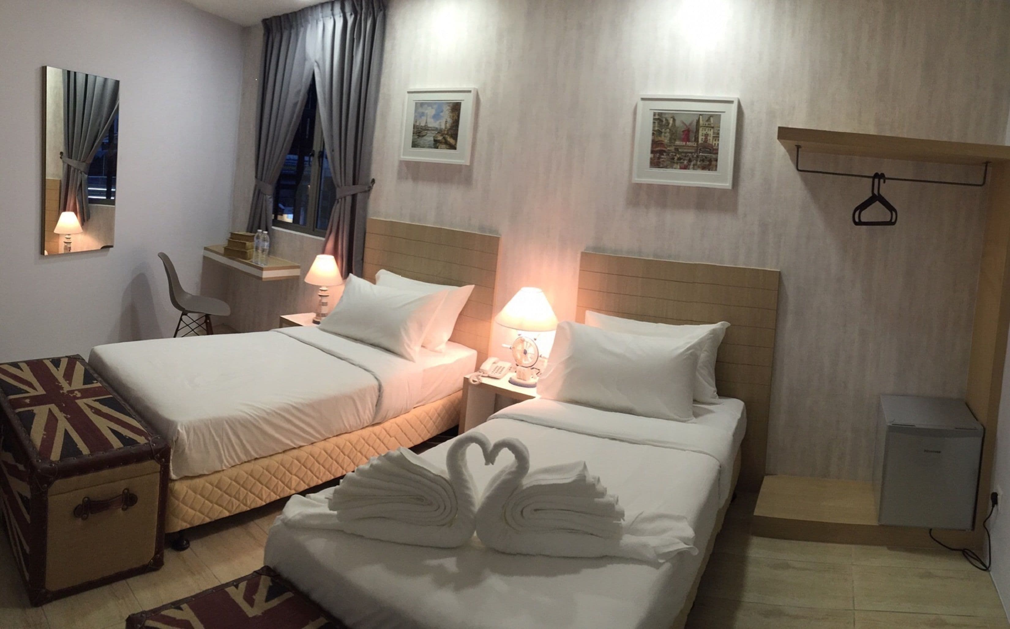 A hotel room with two beds, a small desk, a chair, a bedside table with a lamp, and a wall-mounted TV.