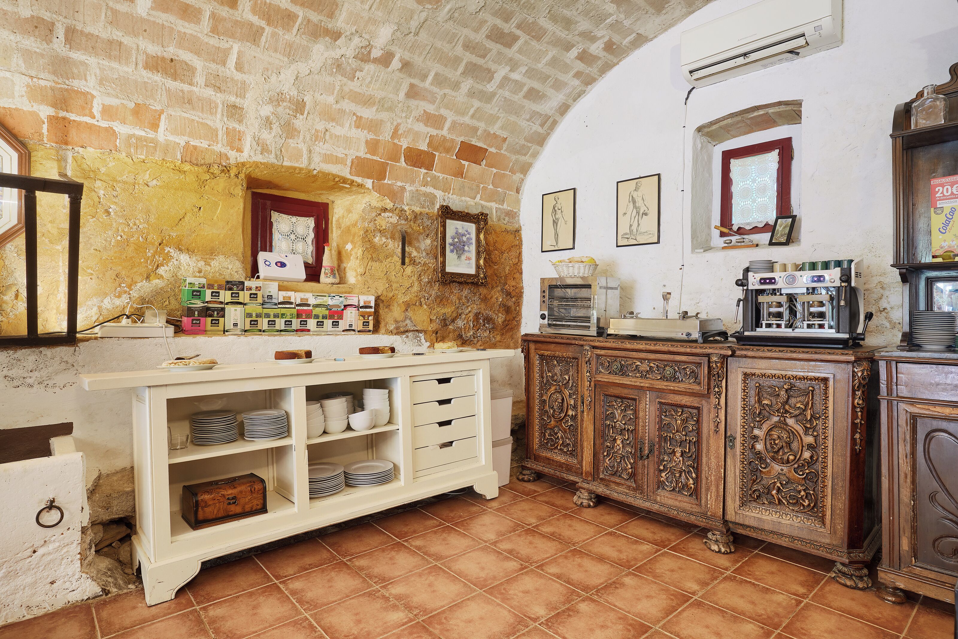 Una cocina con pared de ladrillo, un armario blanco con vajilla y un armario de madera con una cafetera.