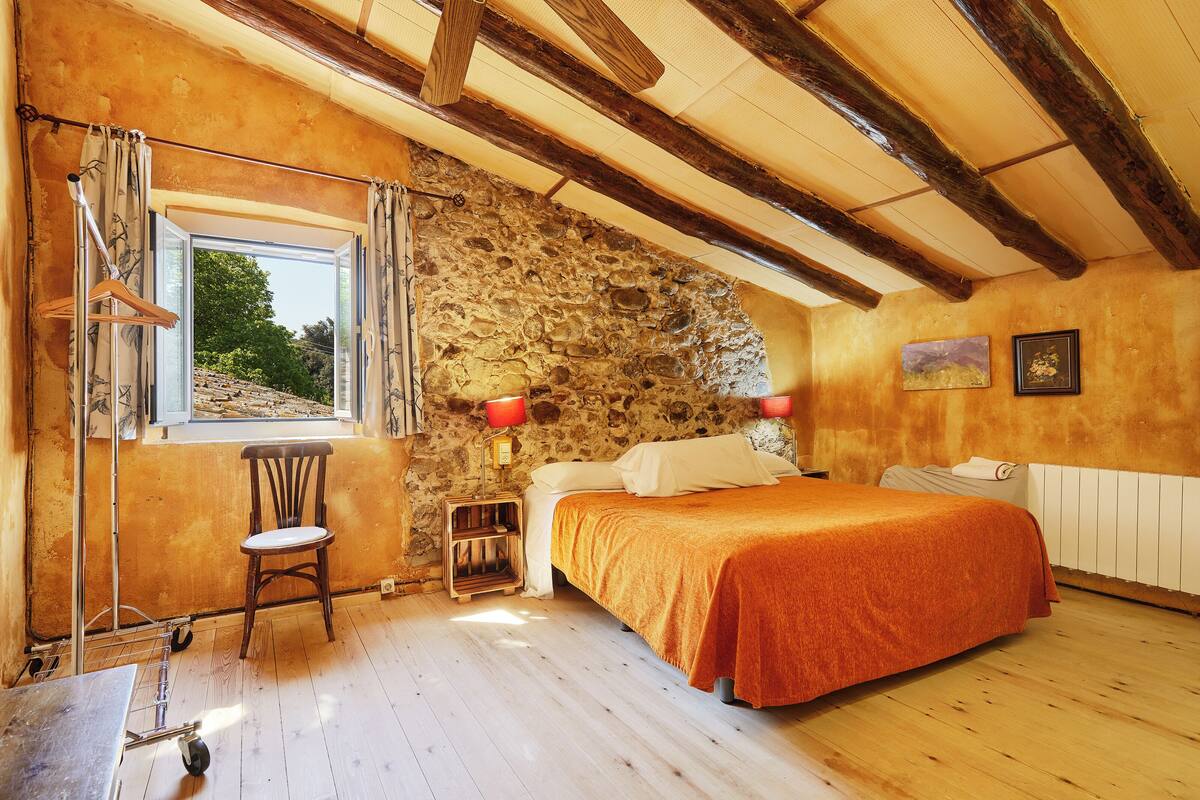 A bedroom with a stone wall, wooden beams, a bed with an orange bedspread, a chair, a window with curtains, and a nightstand with a lamp.