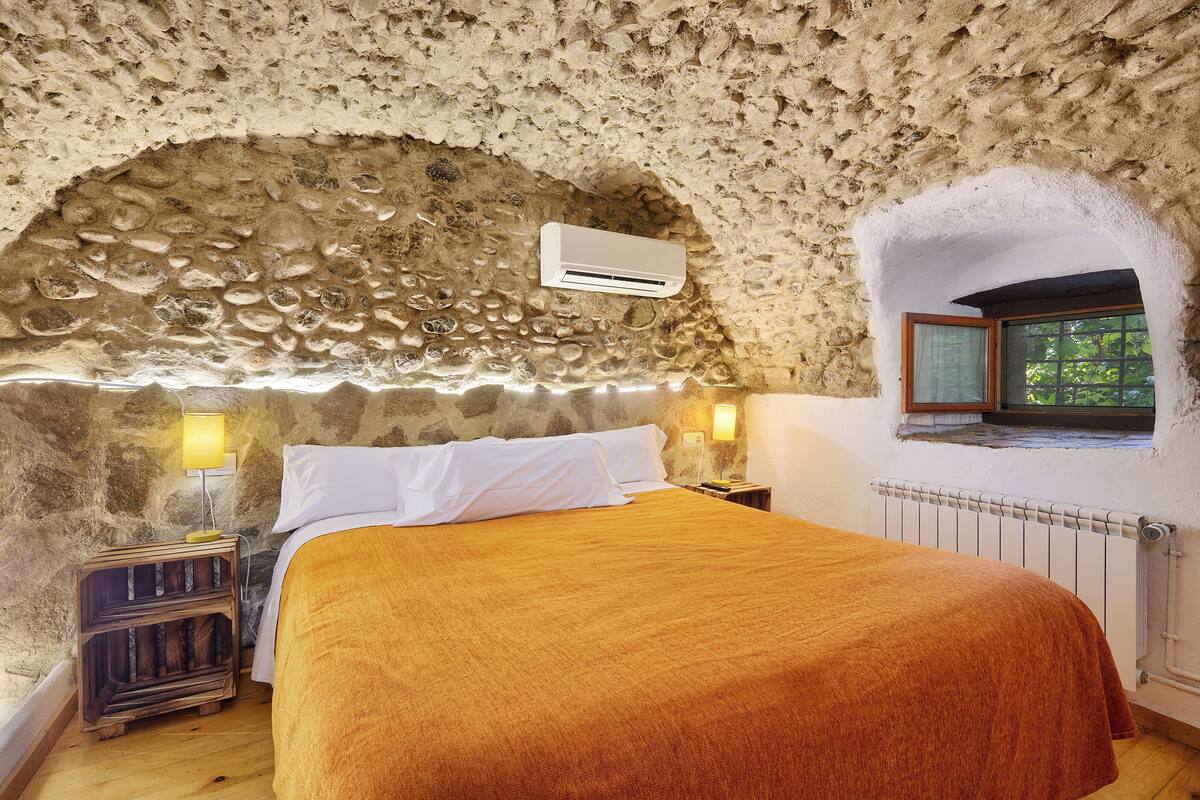 A bedroom with a stone wall, a bed with white and orange bedding, a nightstand with a lamp, and a window with a view of greenery.