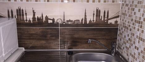 A kitchen with a stainless steel sink, a drying rack, and a tiled backsplash featuring a cityscape design.
